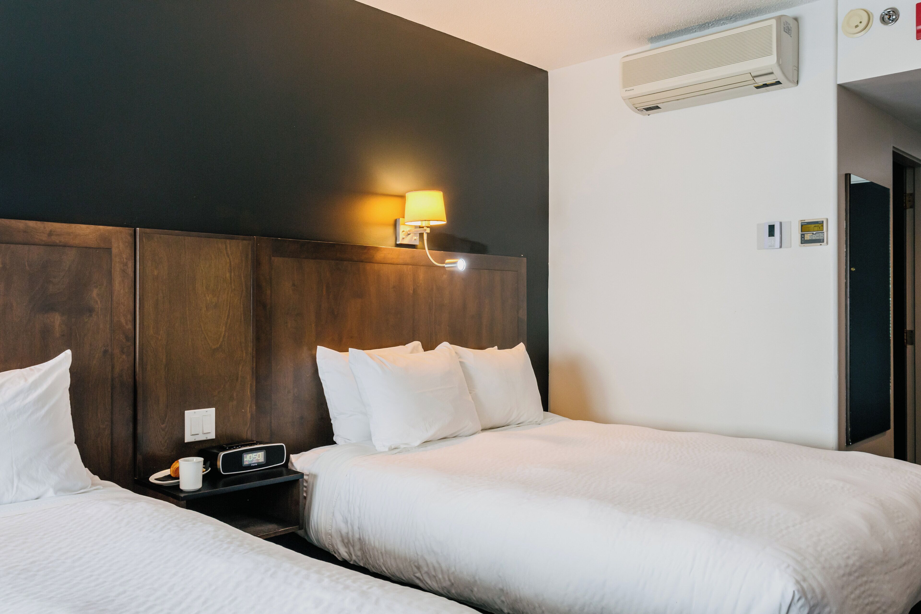 A hotel room with two beds, a nightstand with a clock and a cup, a wall-mounted lamp, and an air conditioning unit.
