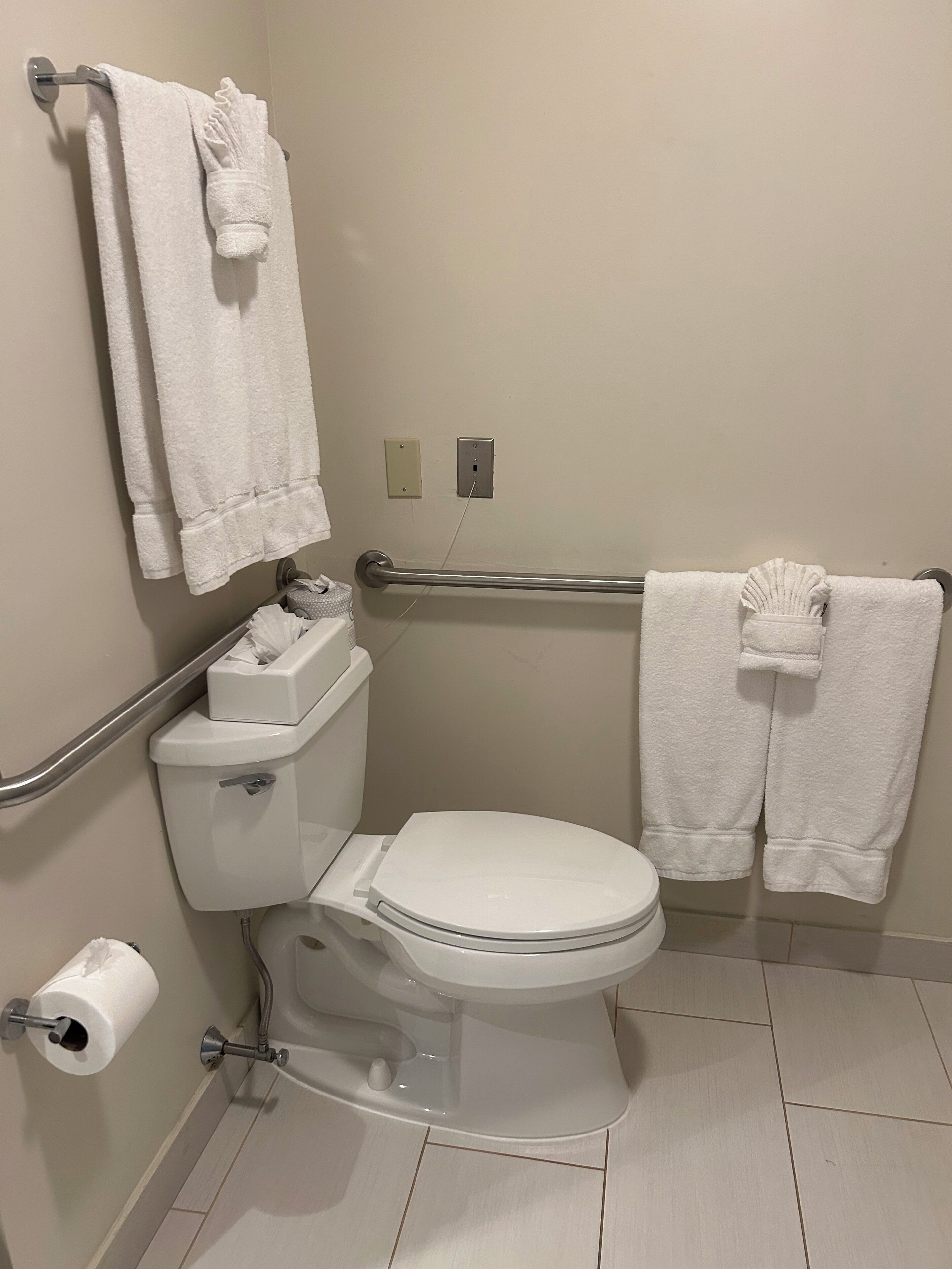 A standard bathroom with a toilet, handrails, a towel rack, and a toilet paper holder.