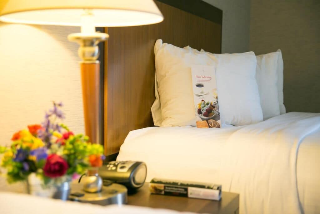 A hotel room with a bed, a nightstand with a lamp, a remote control, a book, and a floral arrangement.