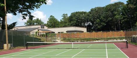 Una cancha de tenis con red, rodeada de árboles y un edificio al fondo.