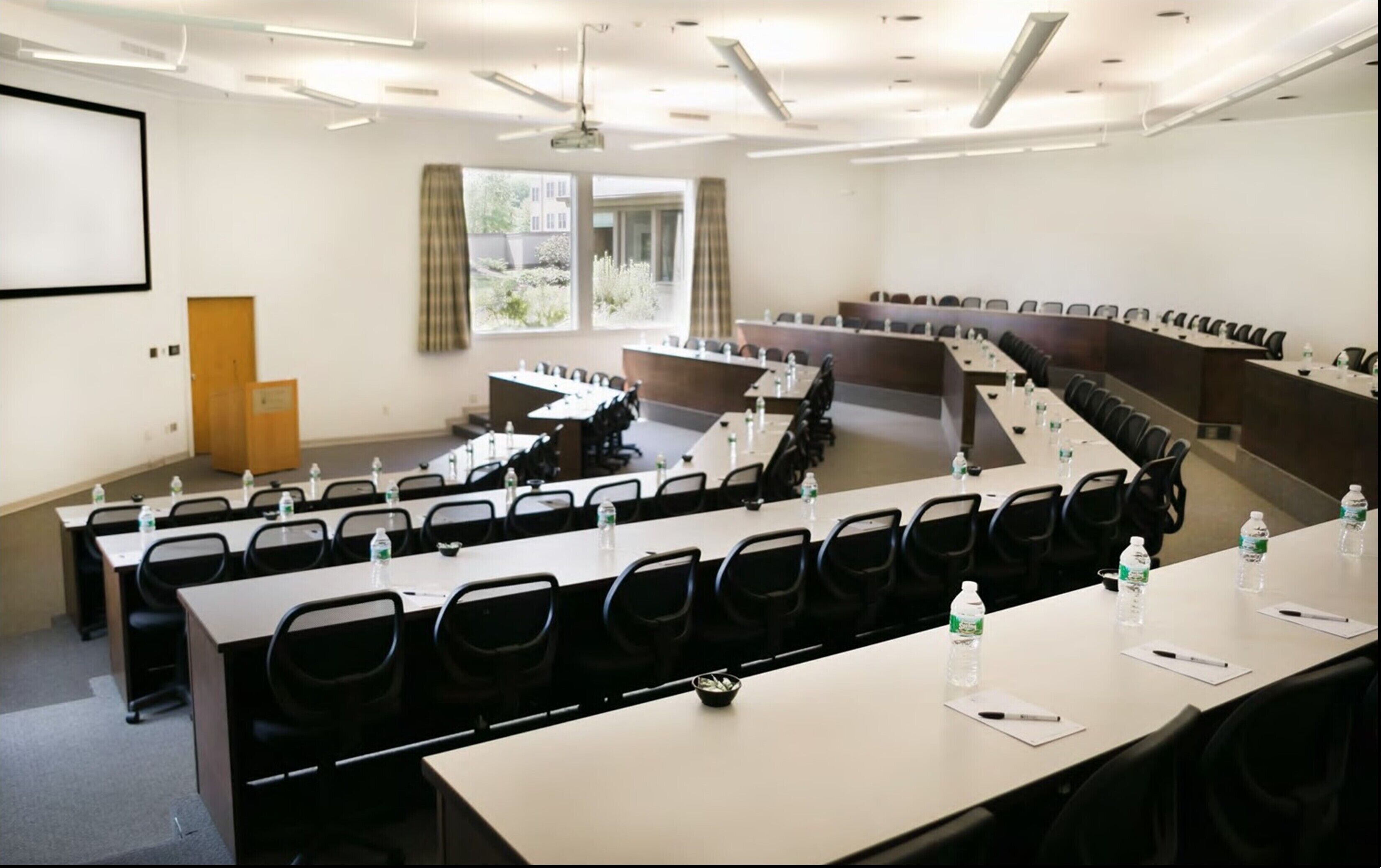 A conference room with multiple tables and chairs, a projector screen, and a window with curtains.