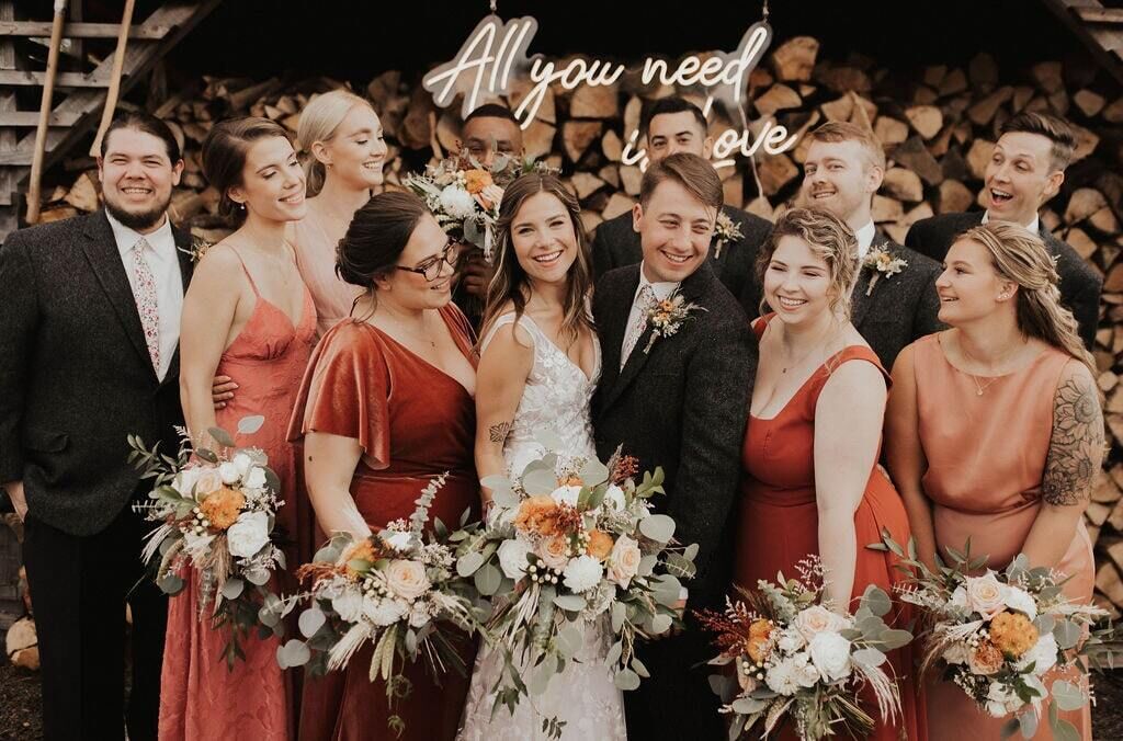A group of people at an outdoor event with a backdrop of stacked logs and a sign that reads "All you need is love."