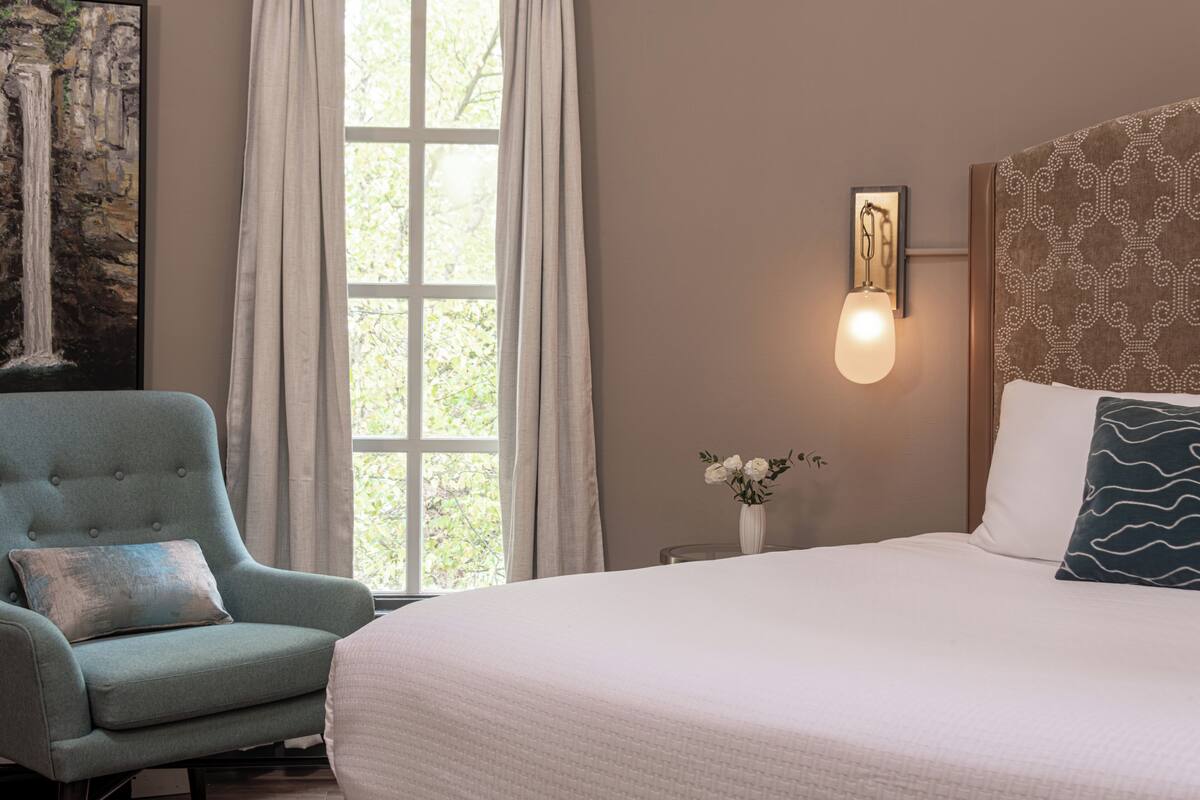 A hotel room with a bed, a chair, a wall-mounted light, a painting, and a window with curtains.