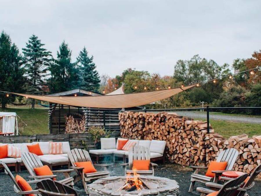 An outdoor seating area with lounge chairs and a fire pit, surrounded by stacked firewood and a covered structure.