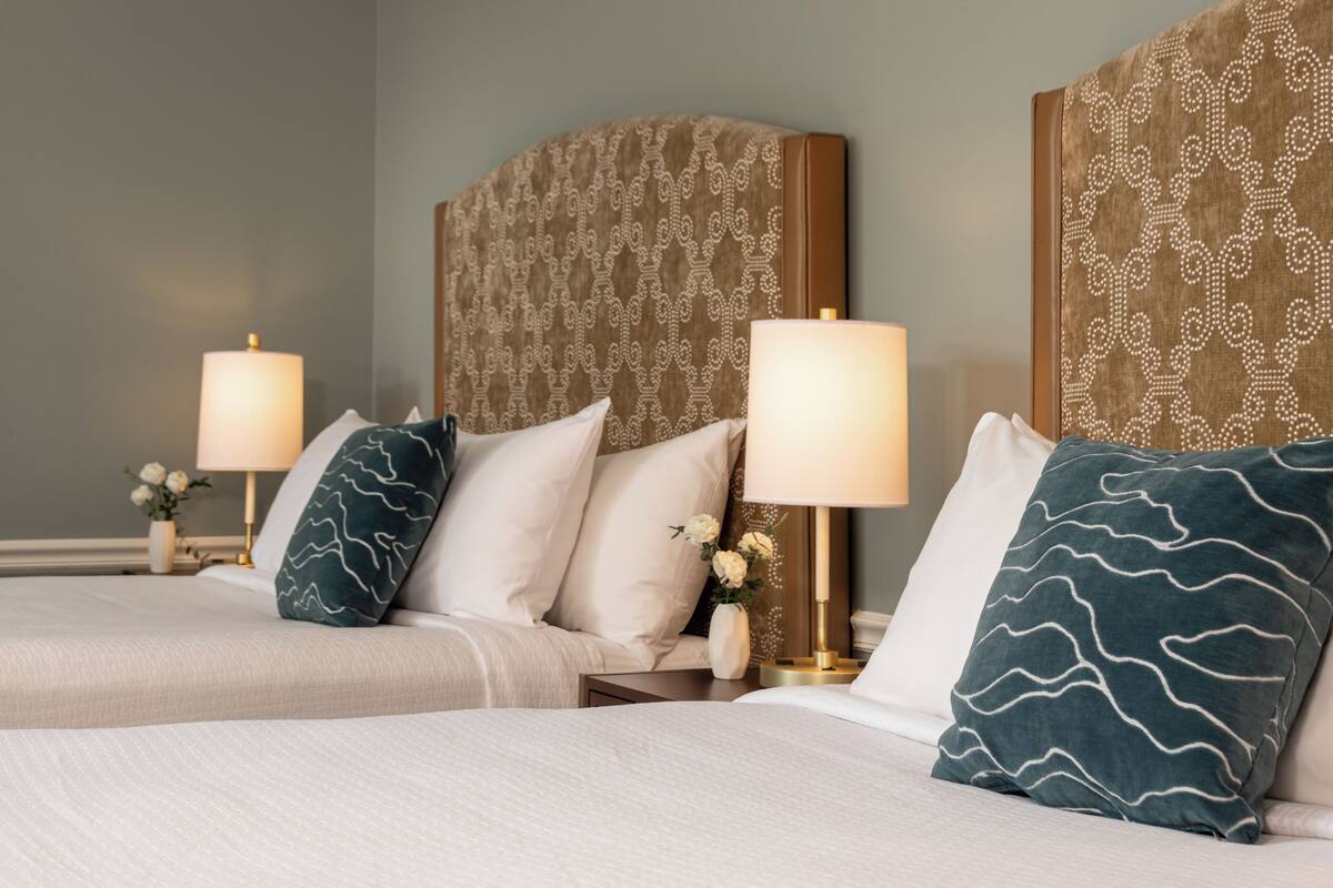 A hotel room with two beds, each with a headboard, bedside tables, lamps, and decorative pillows.