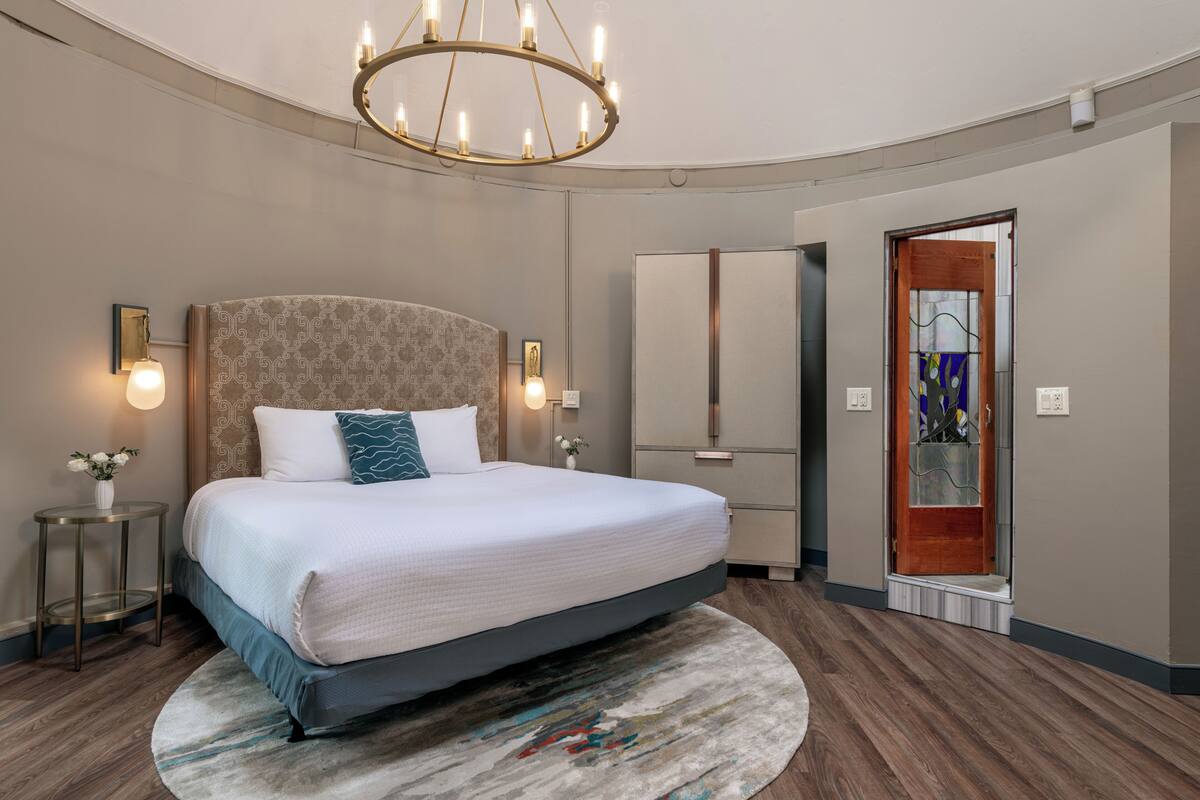 A modern hotel room with a large bed, a round rug, a chandelier, and a wardrobe.