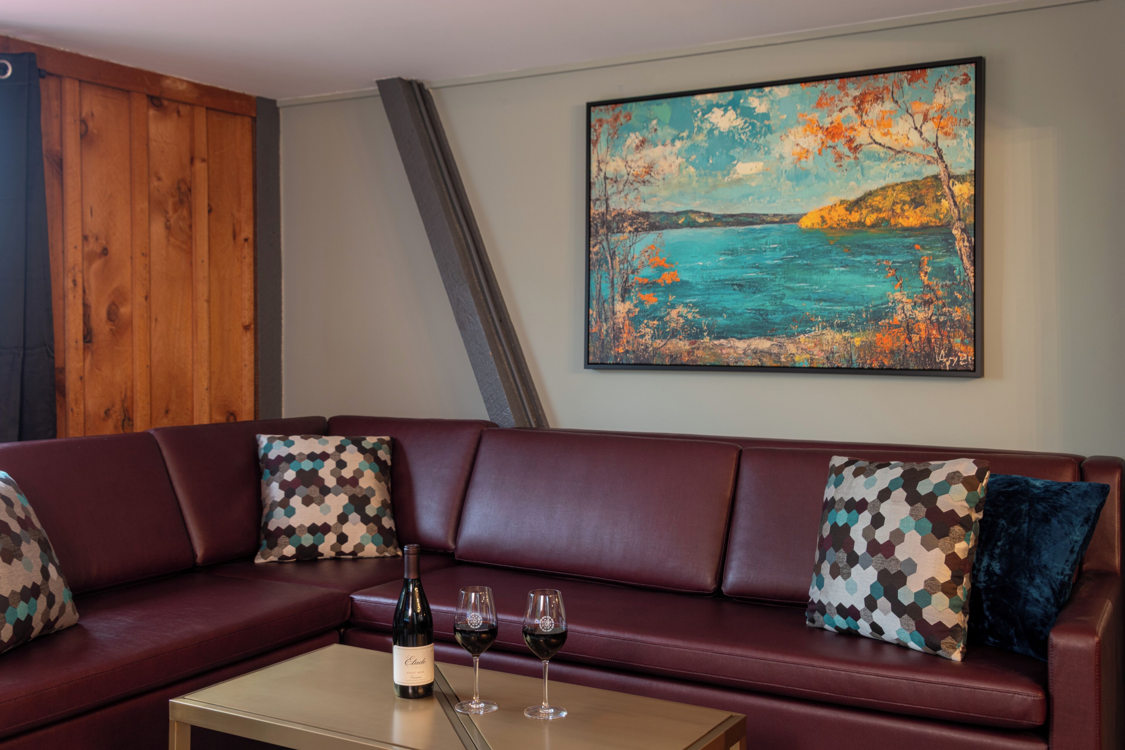 Un canapé en cuir rouge agrémenté de coussins à motifs, une petite table avec une bouteille et des verres, et un tableau représentant un paysage côtier accroché au mur.