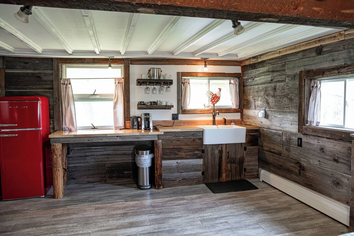 A rustic kitchen with wooden walls, a red refrigerator, a sink, and a window with curtains.