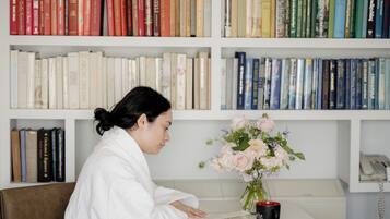 A person in a white robe sitting at a table with a book and a vase of flowers.