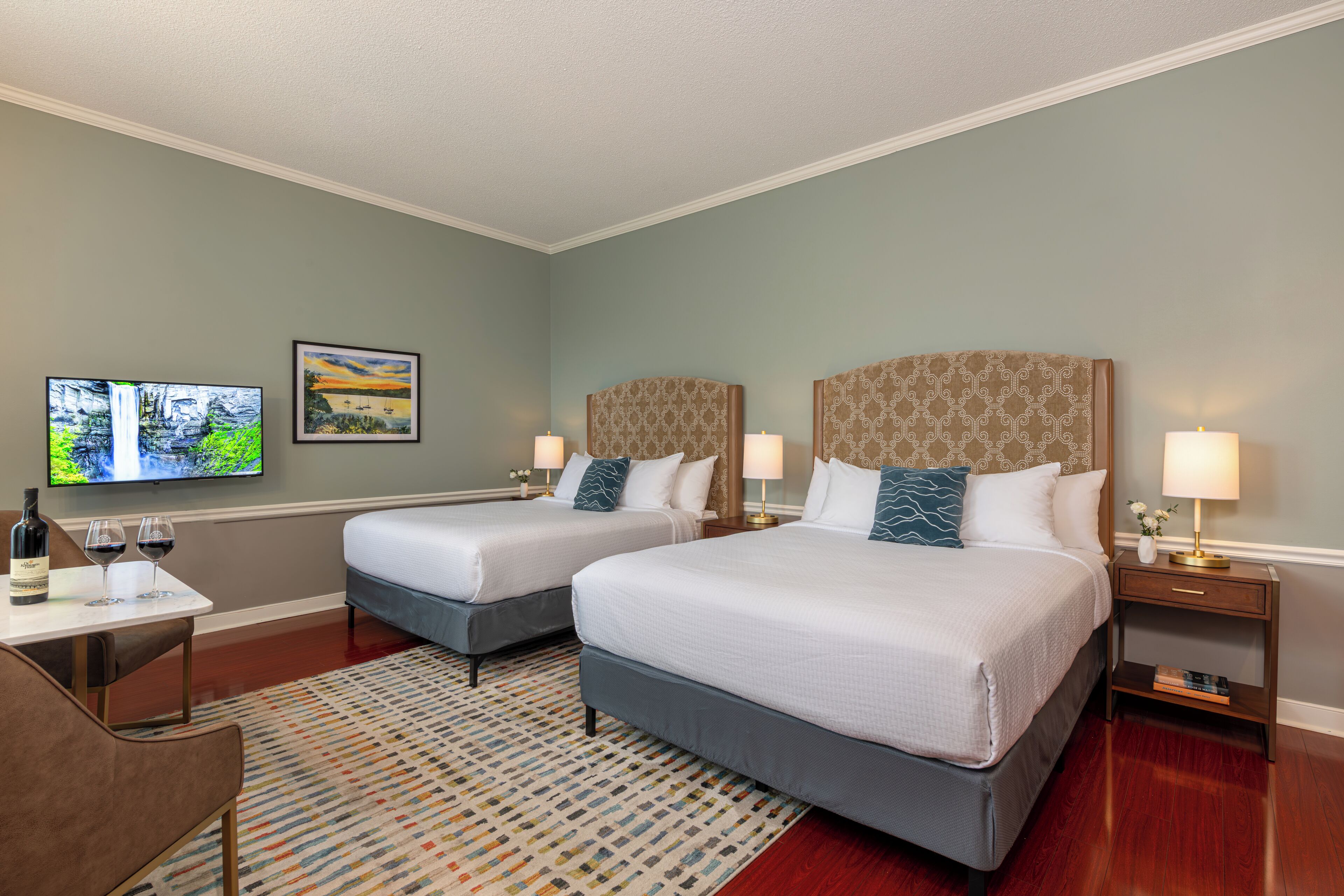 A hotel room with two beds, a TV, a small table with wine glasses, and a patterned rug.