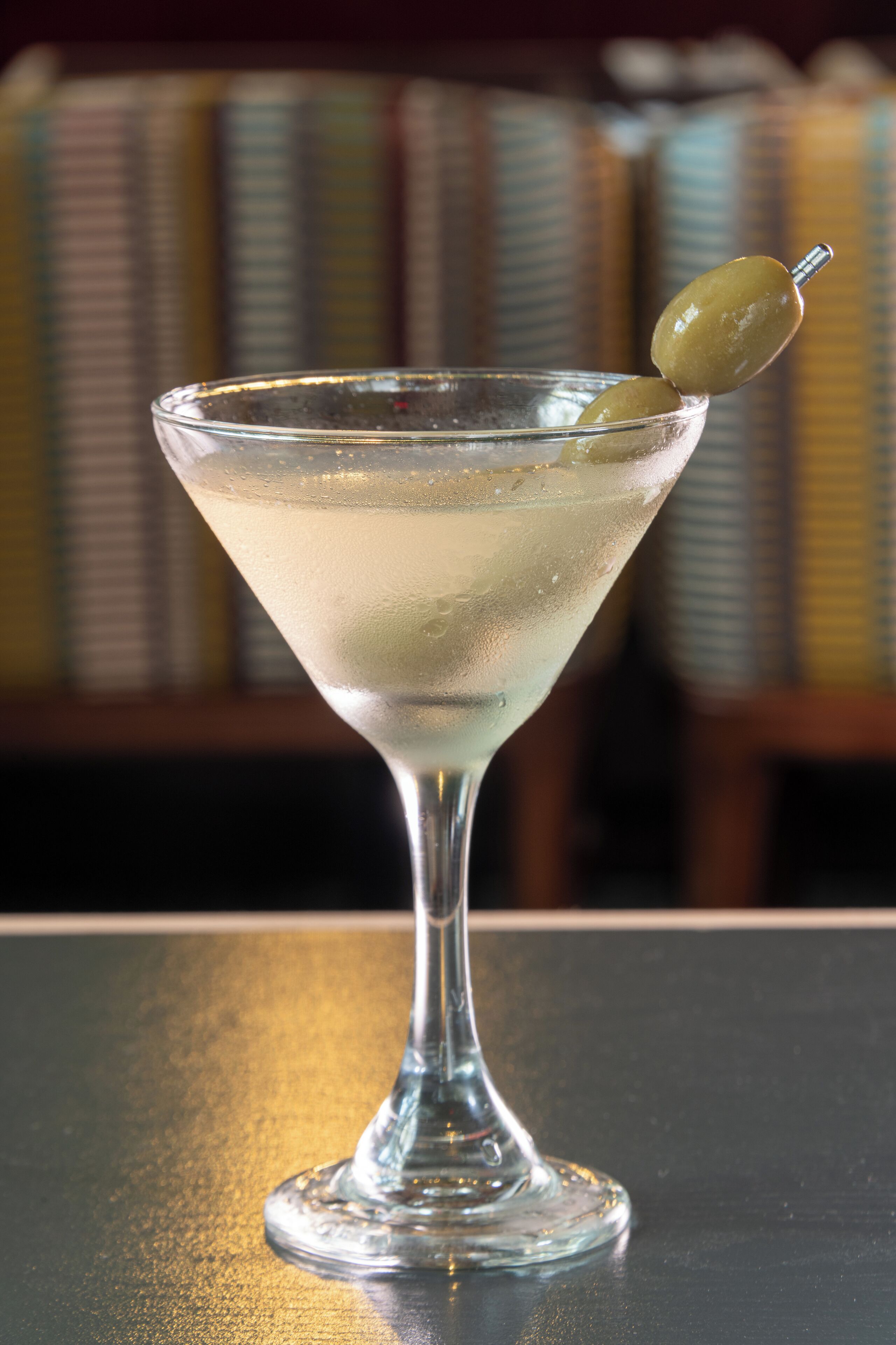 A martini glass with a green olive on the rim, placed on a table.