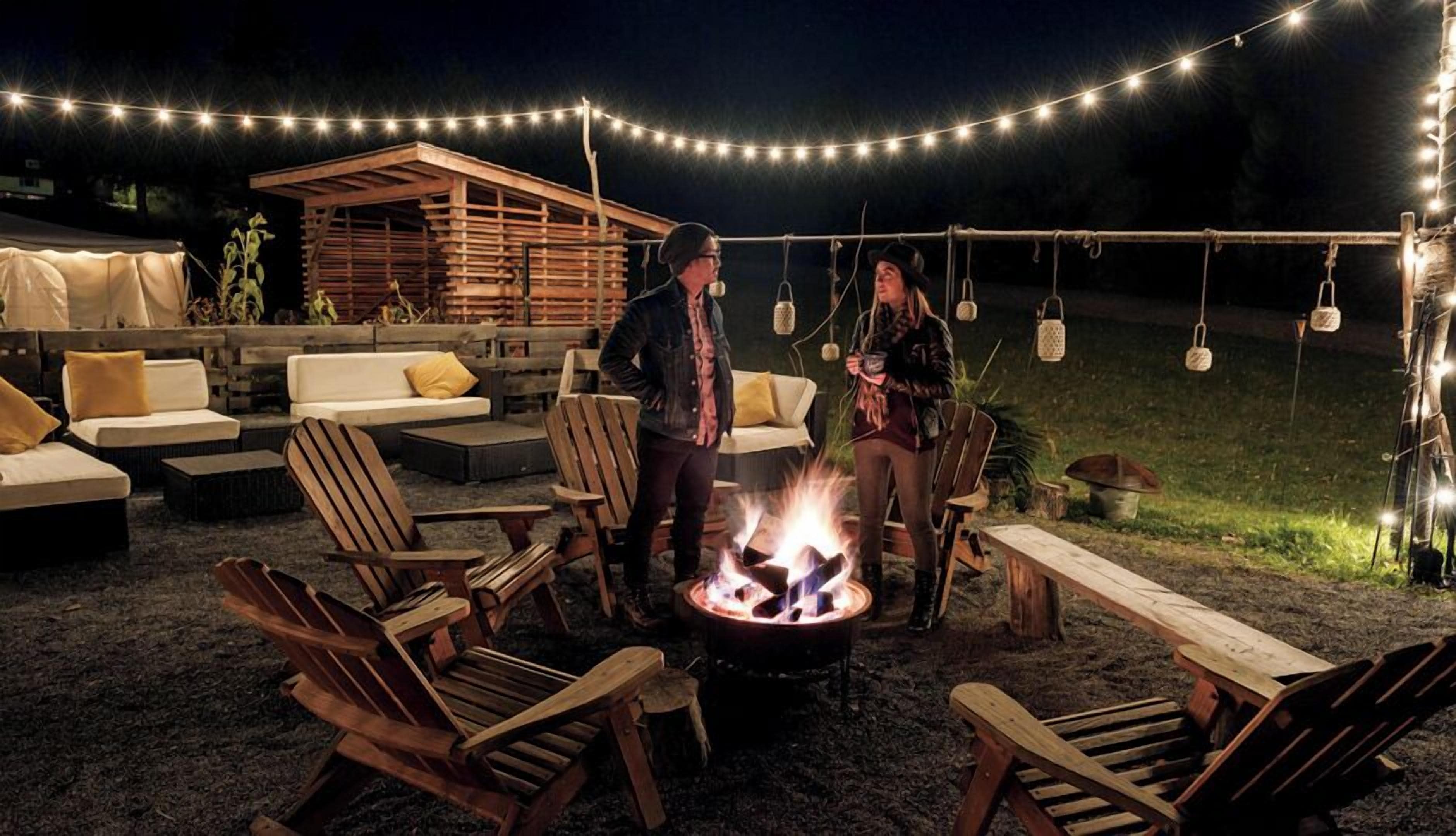 Un espacio al aire libre con sillas de madera y un brasero, rodeado de luces colgantes y una estructura de madera al fondo.