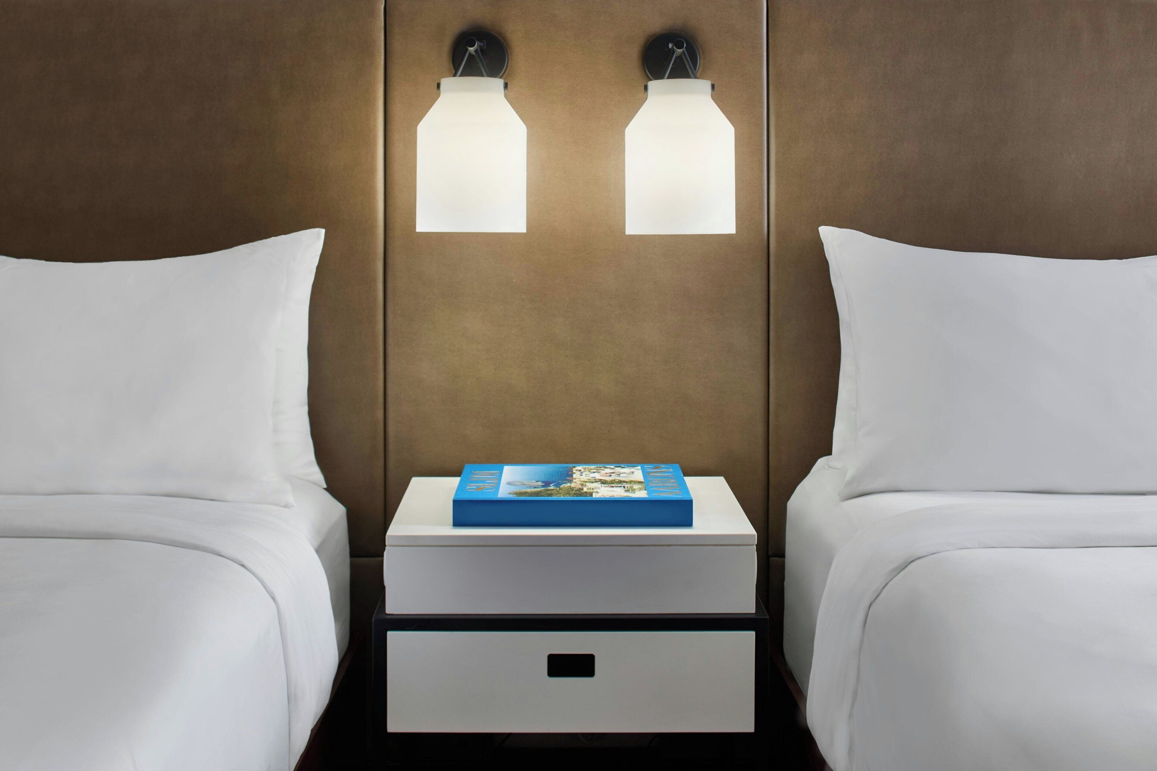 A hotel room with two beds, a nightstand with a book, and wall-mounted lights.