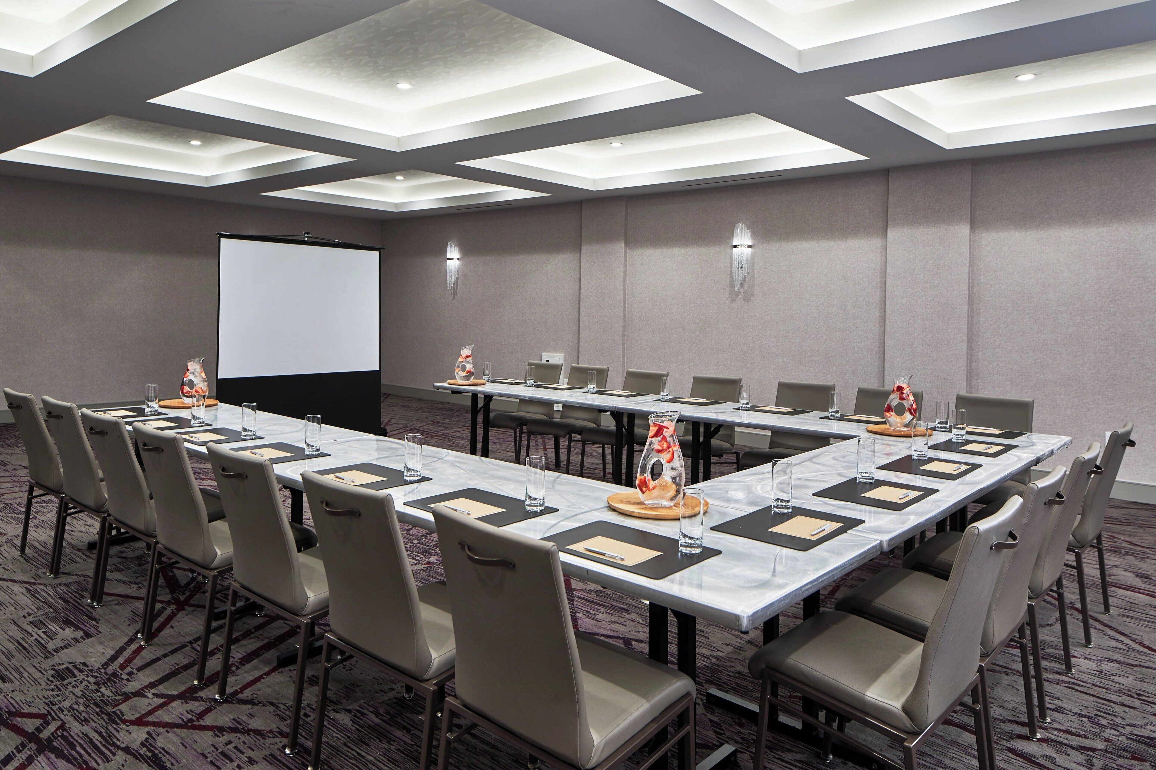 Un salón de conferencias con una larga mesa, sillas, una pantalla de proyector y piso alfombrado.