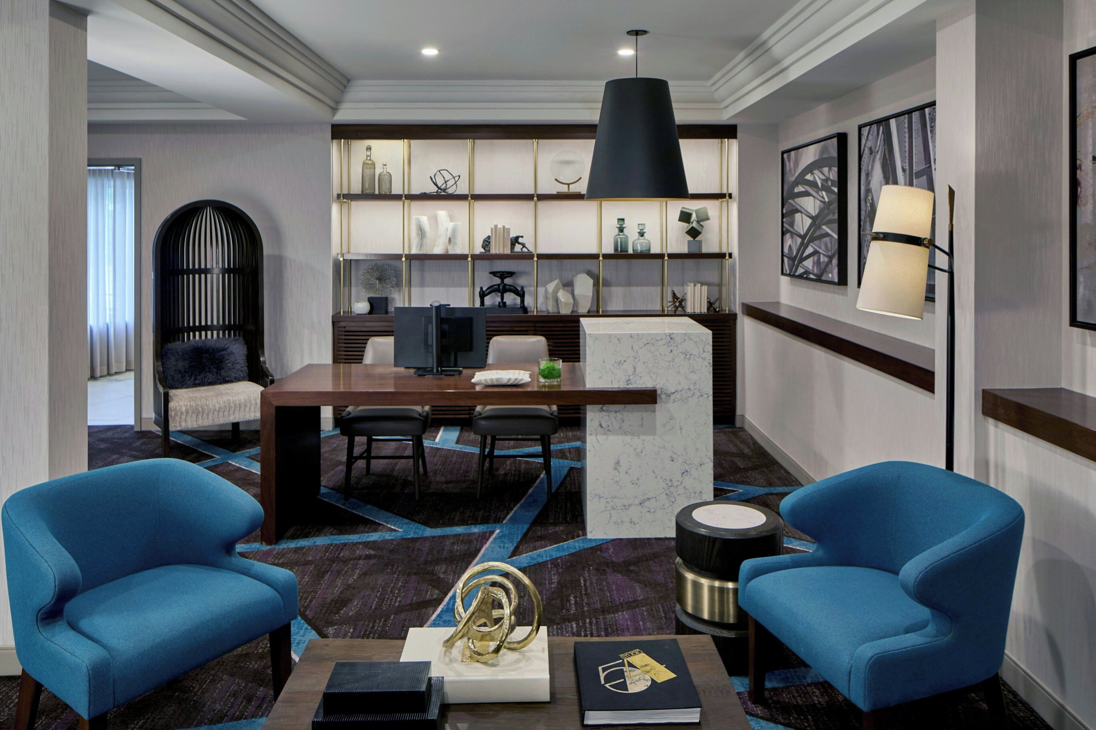Een hotellobby met een receptiebalie, twee blauwe fauteuils, een marmeren salontafel, een boekenplank met decoratieve objecten, en een grote zwarte hanglamp.