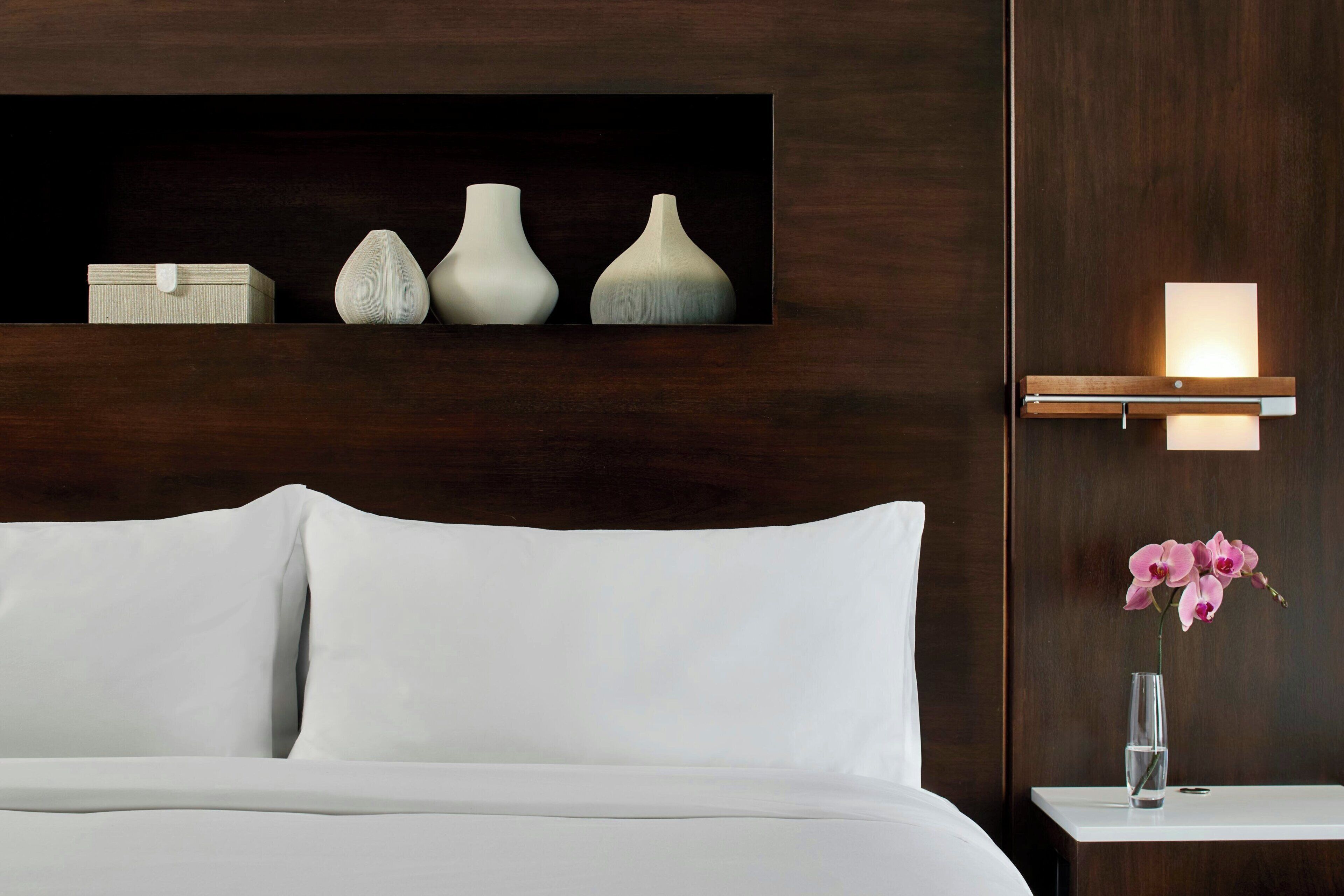 A hotel room with a bed, a bedside table with a vase of flowers, a wall-mounted light, and a shelf with decorative vases.