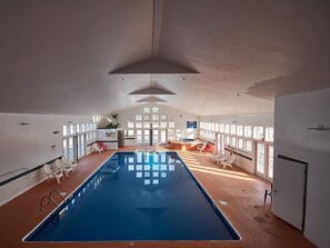 An indoor swimming pool with a long, narrow shape, surrounded by white chairs and a sunlit area with a hot tub.