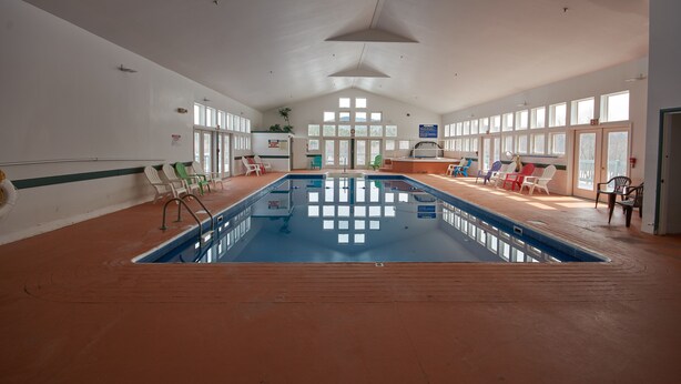 An indoor swimming pool with a wooden deck, multiple lounge chairs, and large windows allowing natural light. - River View Resort (Bethel)