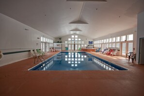 Una piscina interior con cubierta de madera, múltiples sillas de descanso y grandes ventanales que dejan entrar luz natural.