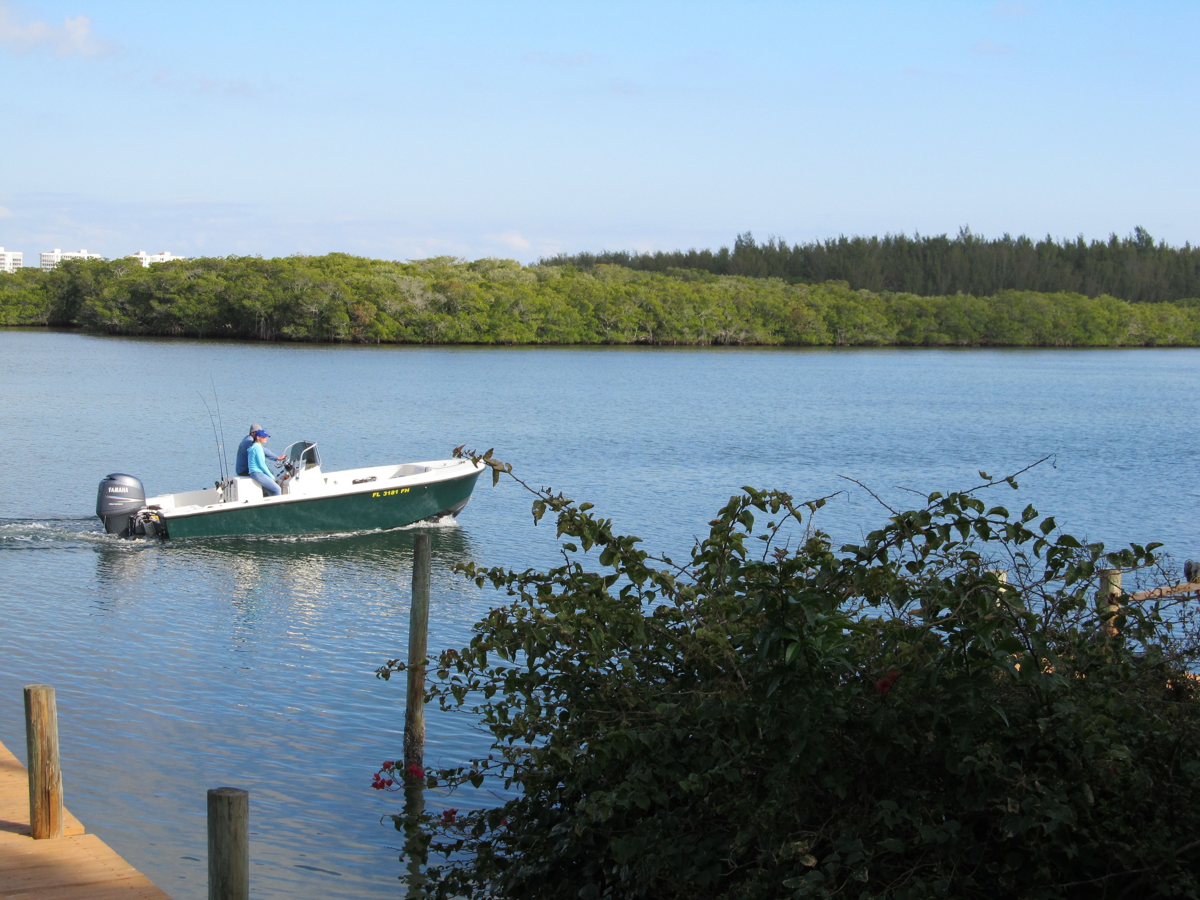 A boat on calm water with a dock in the foreground and a forested shoreline in the background.