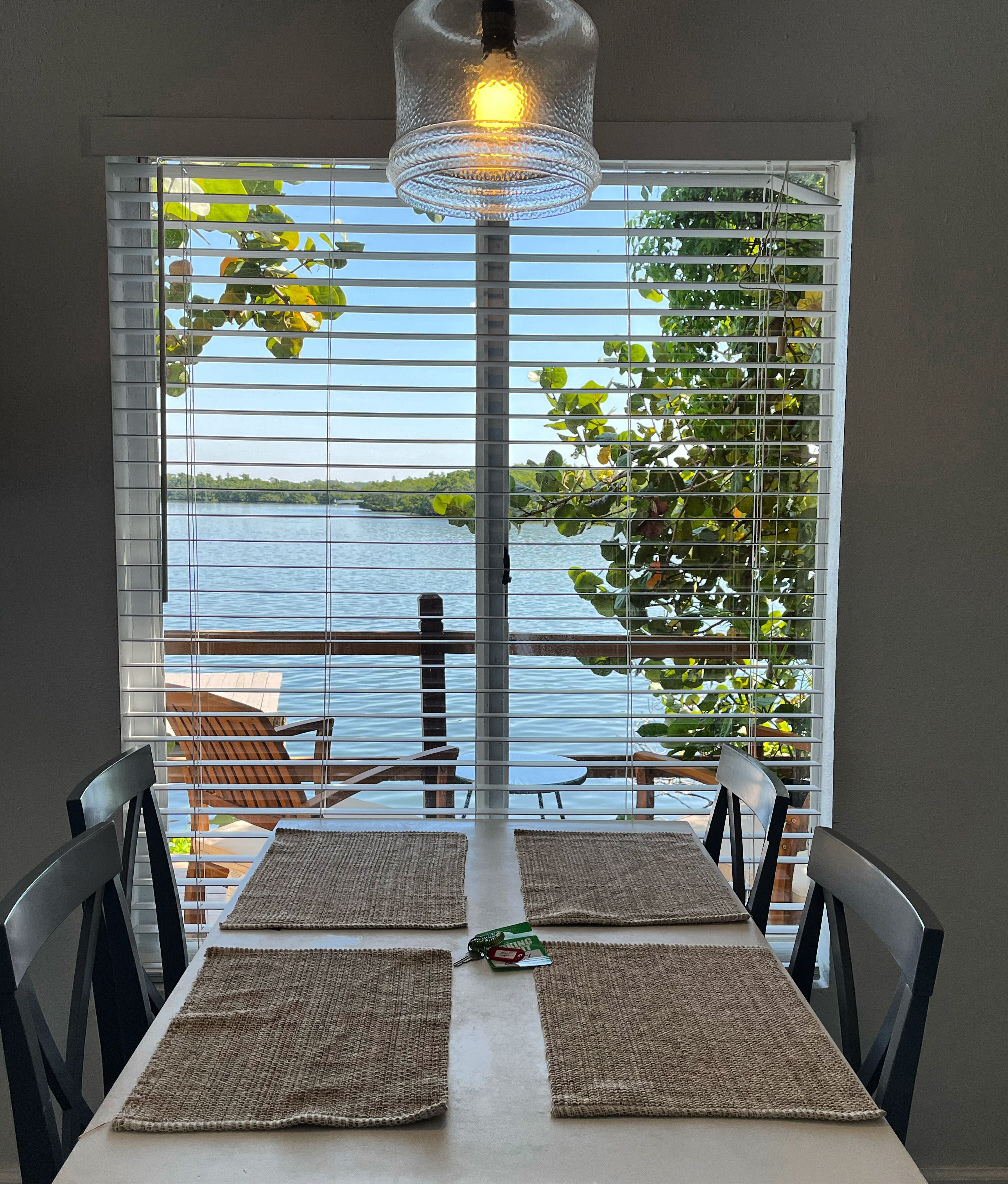 A dining area with a table set for two, a hanging light fixture, and a view of a body of water through window blinds.
