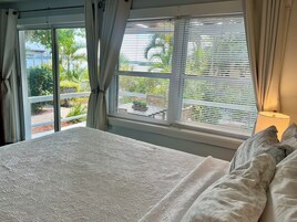 Quarto com cama, janela com persianas e vista para o verde lá fora.