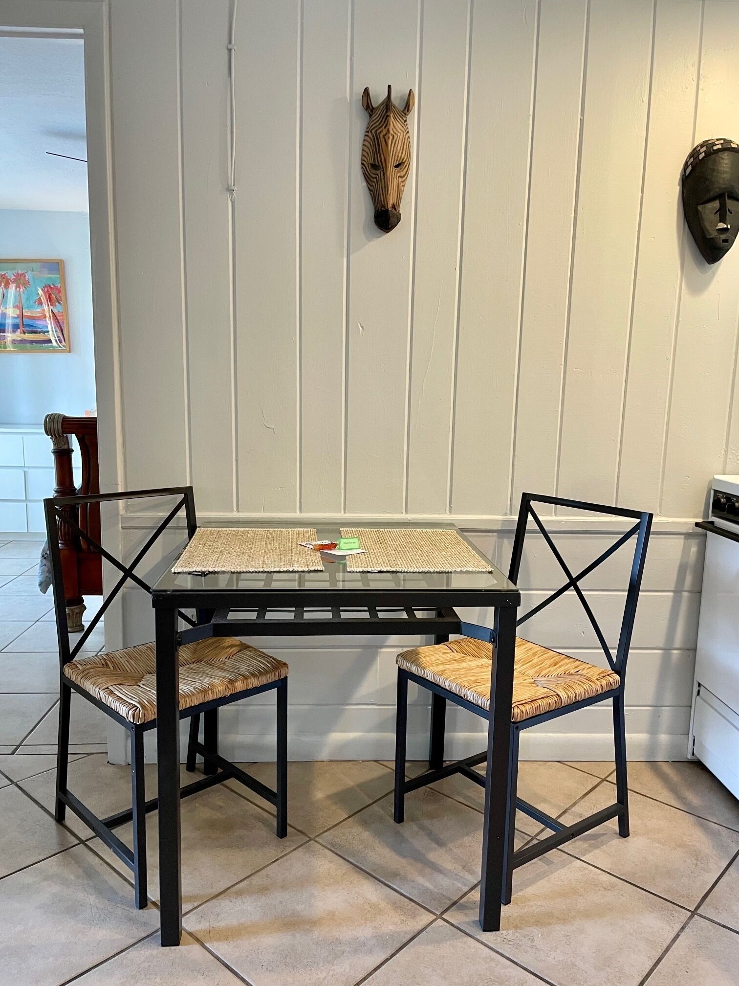 Un comedor con una mesa de vidrio, dos sillas con asientos de mimbre y una escultura de cabeza de cebra montada en la pared.