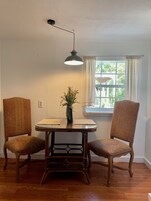 Un comedor con una mesa redonda, dos sillas, una lámpara colgante, una ventana con cortinas y una planta sobre la mesa.