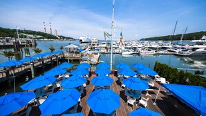 Un puerto deportivo con barcos amarrados, un muelle con sombrillas azules y sillas blancas, y un asta con banderas.