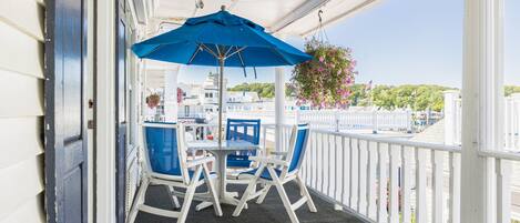 A covered outdoor seating area with a table and chairs, a blue umbrella, and a view of a white building and greenery in the background.