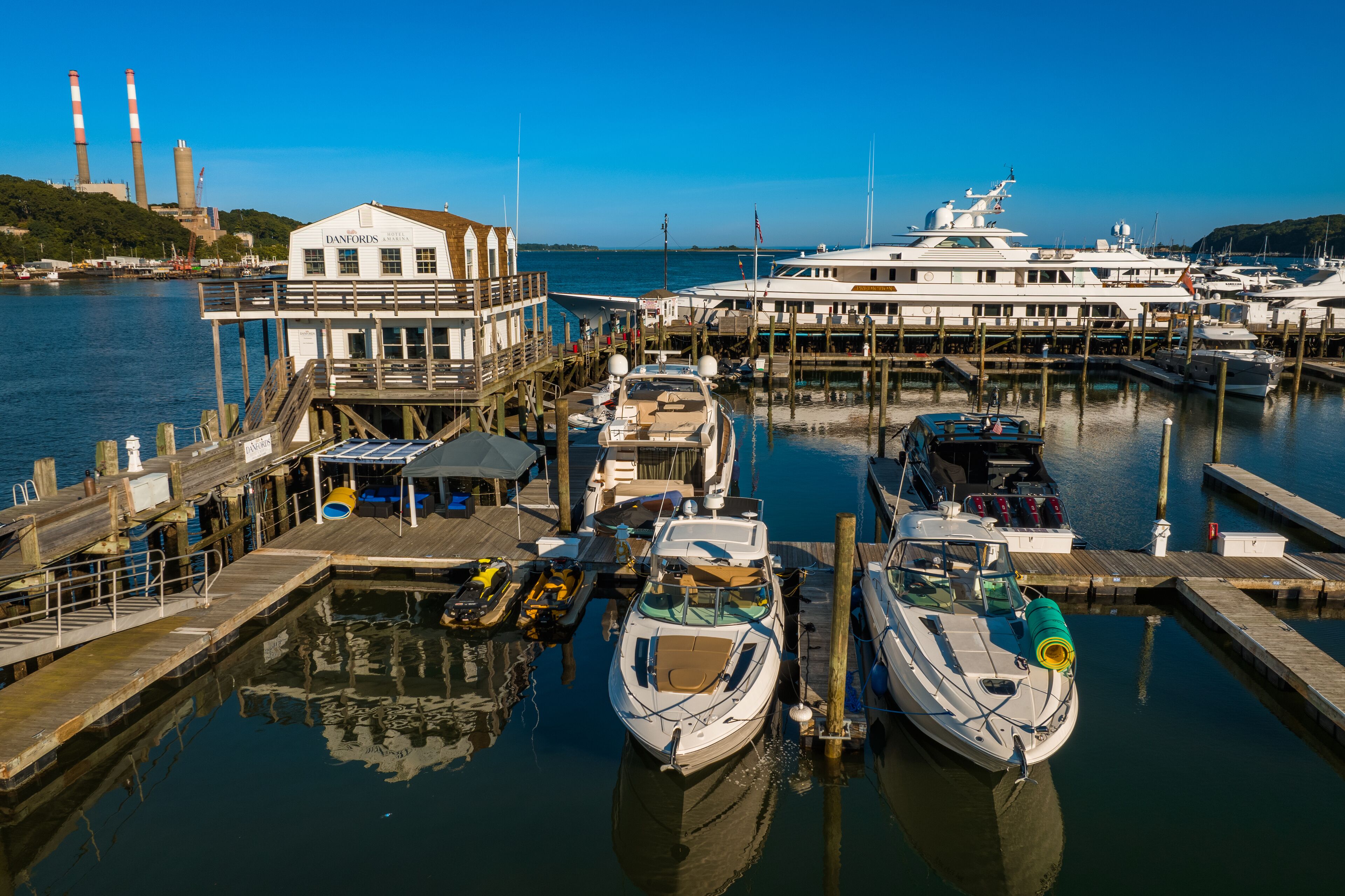 A marina with several boats docked, a building labeled "DANFORTHS," and a distant industrial facility with smokestacks.