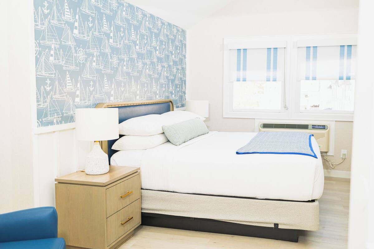 A neatly arranged bedroom with a bed, bedside table, lamp, and a wall with a nautical pattern.