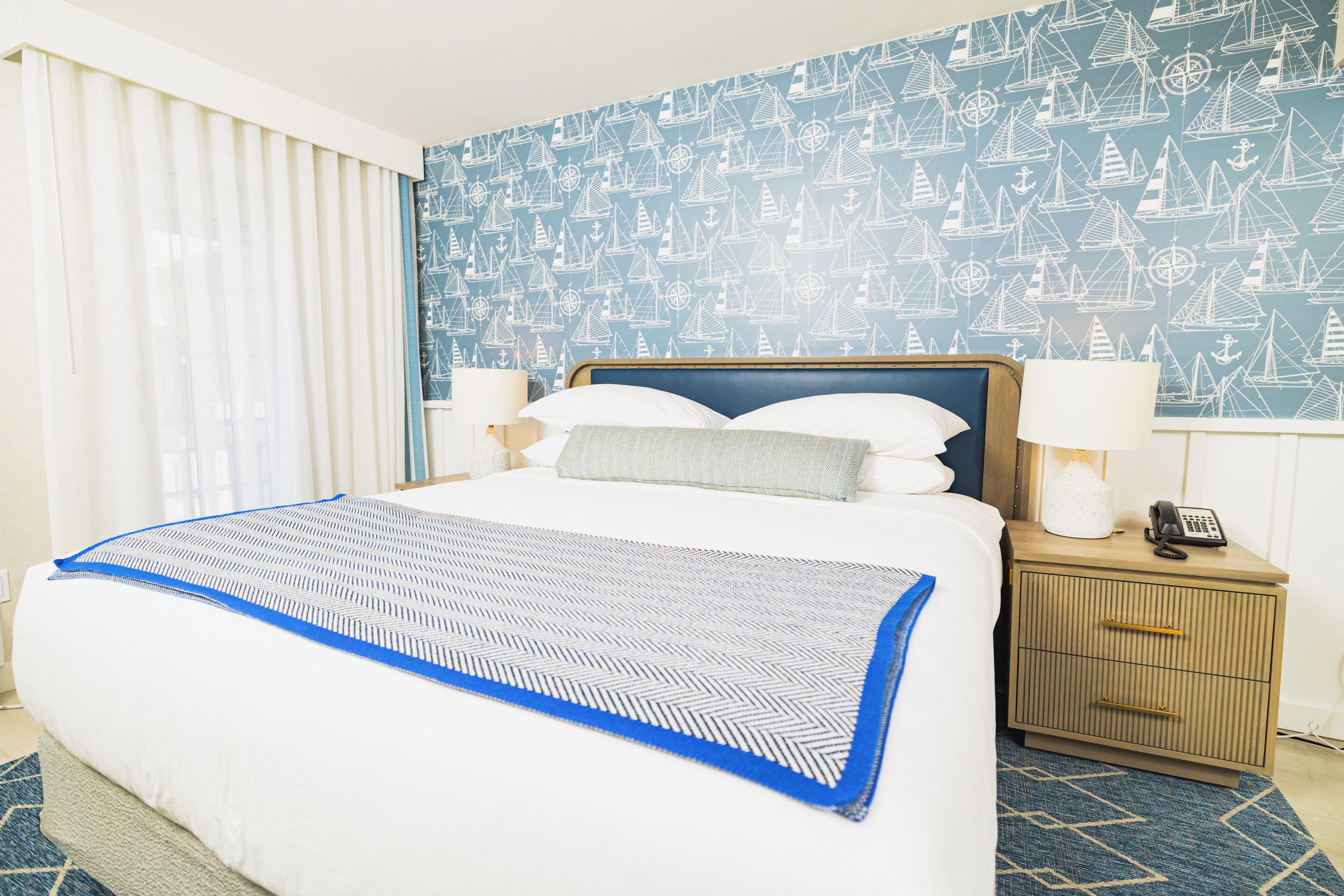 A hotel room with a large bed, a nightstand with a lamp, and a wall with nautical-themed wallpaper.