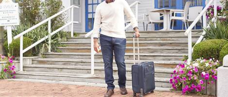 A man with a suitcase standing on a brick pathway in front of a building with blue doors and white railings.