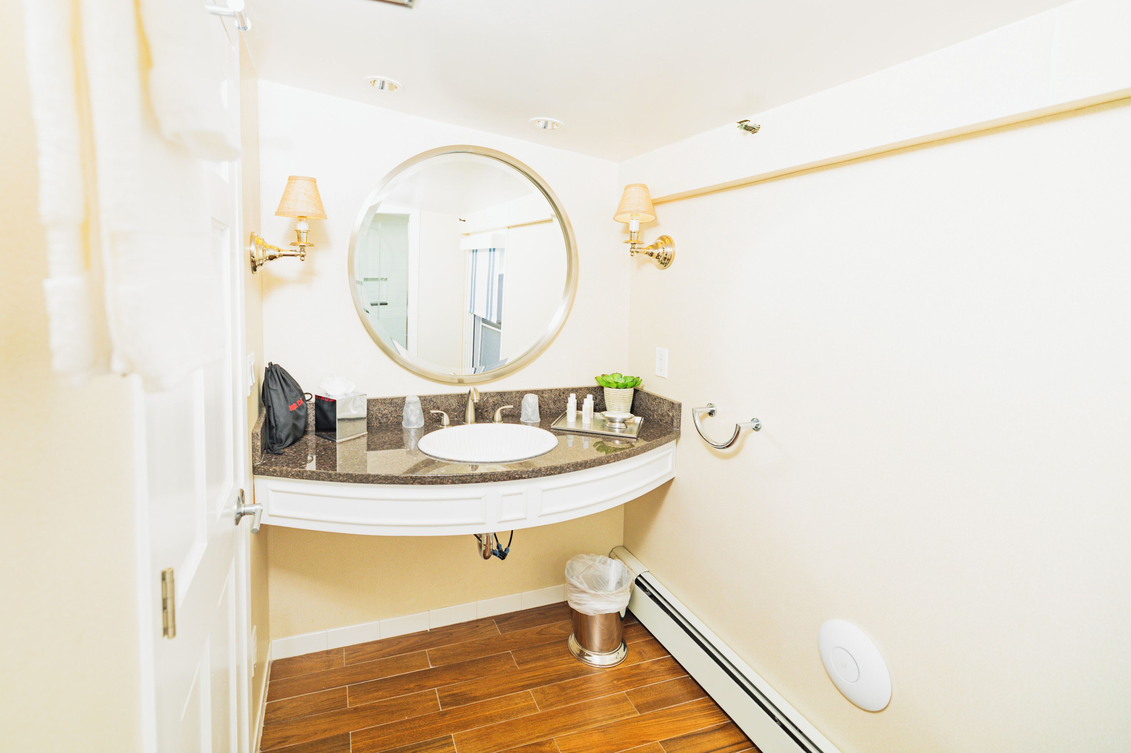 Un baño con un lavabo blanco, un espejo redondo, un perchero y un cubo de basura.