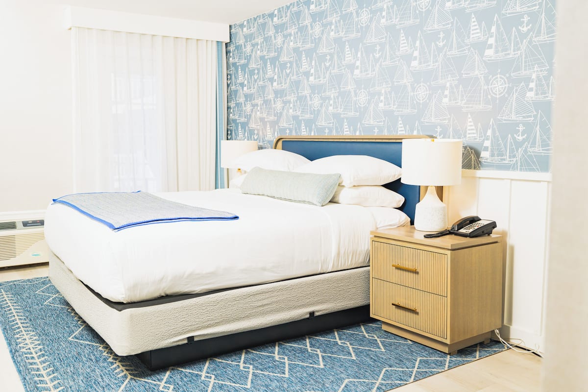 A neatly made bed with pillows and a blanket, a bedside table with a lamp and phone, and a patterned wall.