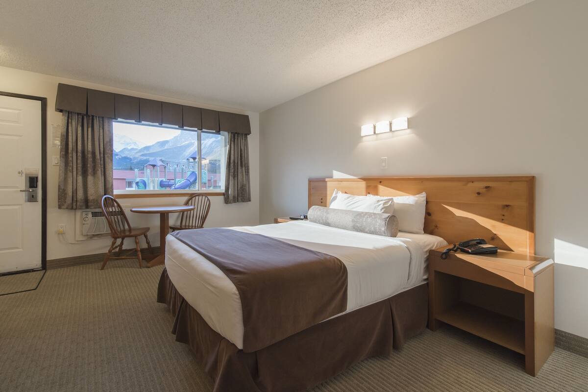 Habitación de hotel con cama, escritorio, silla, mesita de noche y ventana con vista a las montañas.