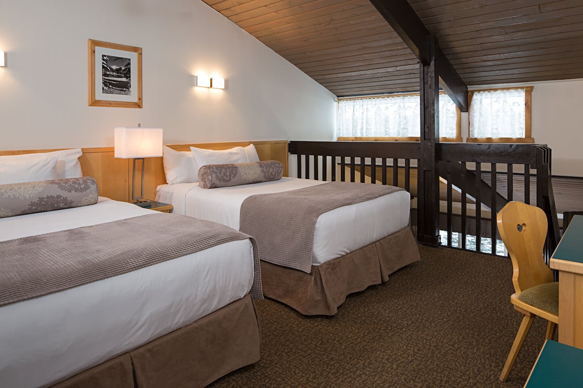 Habitación de hotel con dos camas, un escritorio y una silla. Tiene una ventana con cortinas y una escalera de madera.