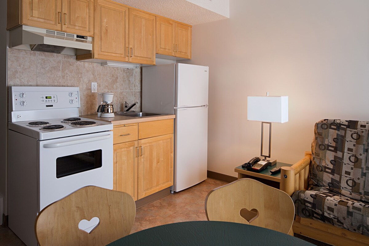 A compact kitchen with a stove, refrigerator, wooden cabinets, and a dining area with two chairs.