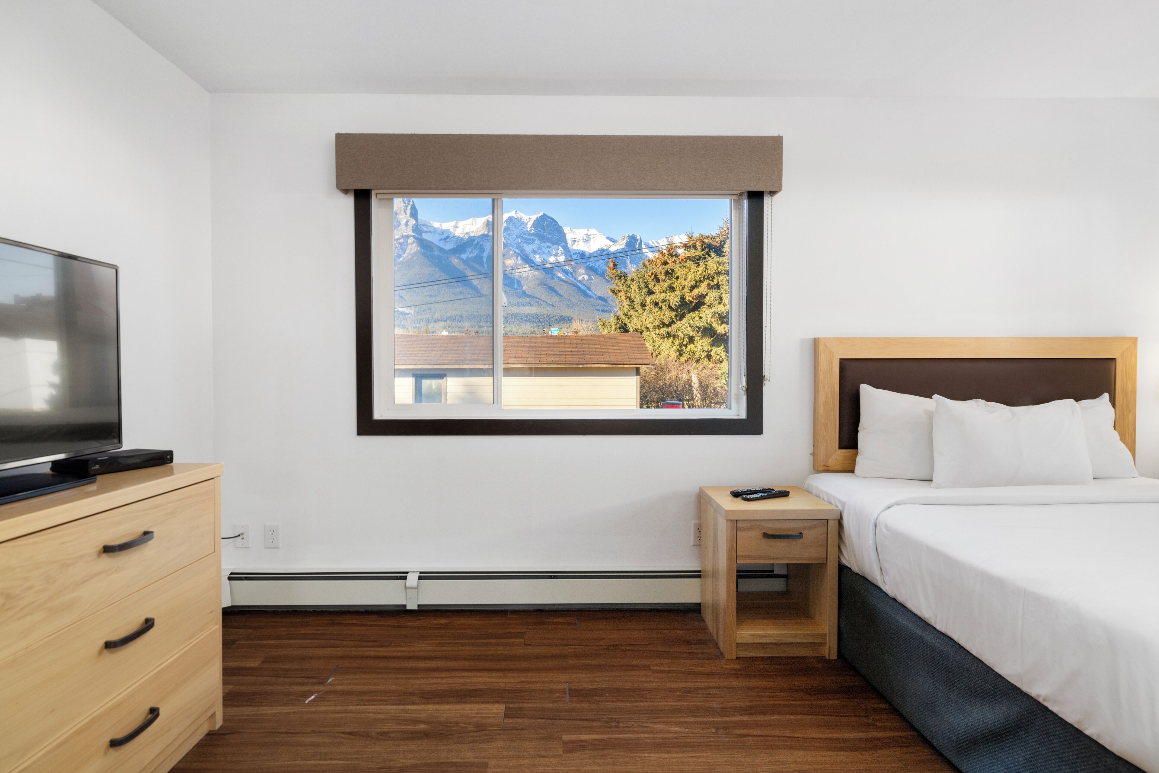 Habitación de hotel con cama, un televisor sobre un aparador de madera, una mesita de noche pequeña y una ventana con vistas a la montaña.