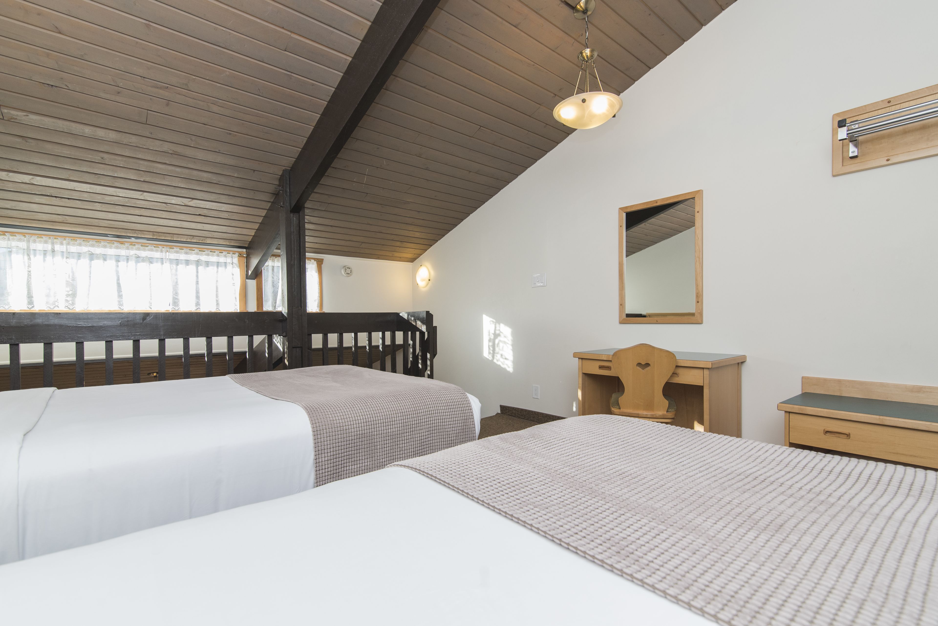 A room with two beds, wooden headboards, a wooden desk, a mirror, and a ceiling with wooden beams.