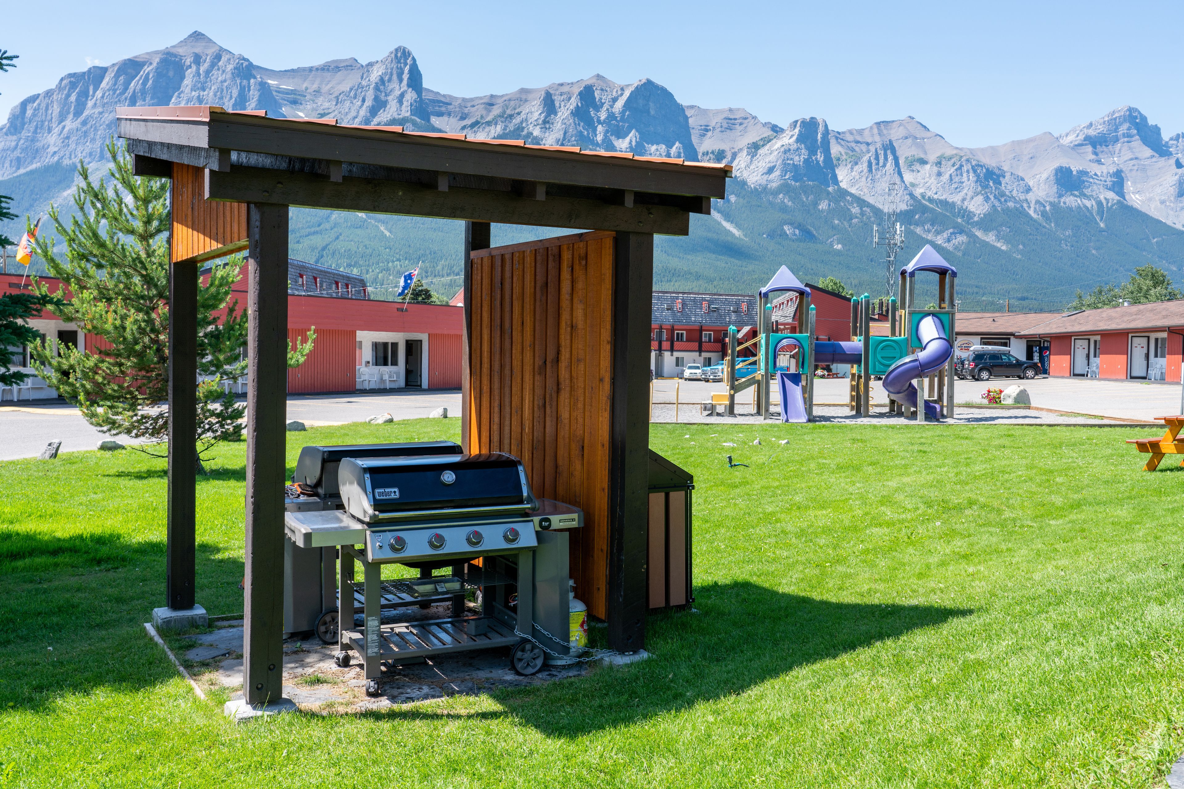Un área de barbacoa con el paisaje de una montaña al fondo, un parque infantil y un edificio con tejado rojo.