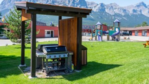 A barbecue area with a mountain backdrop, a playground, and a building with a red roof. - Rocky Mountain Ski Lodge (Canmore)
