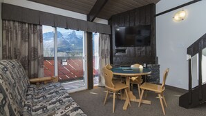 A cozy living room with a sofa, a dining table with chairs, a flat-screen TV, and a view of mountains through large windows. - Rocky Mountain Ski Lodge (Canmore)