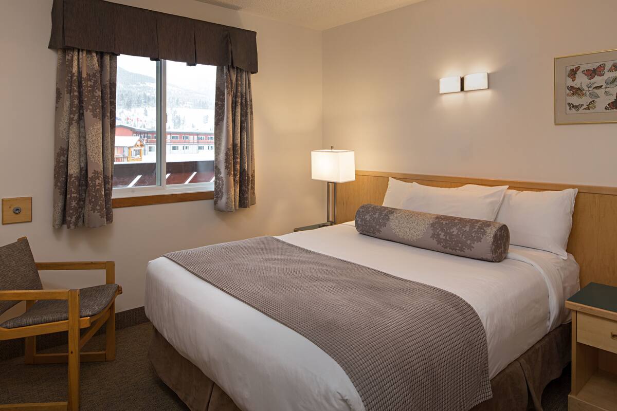 Una habitación de hotel con una cama grande, una silla, una mesita de noche, una lámpara y un ventanal con cortinas.