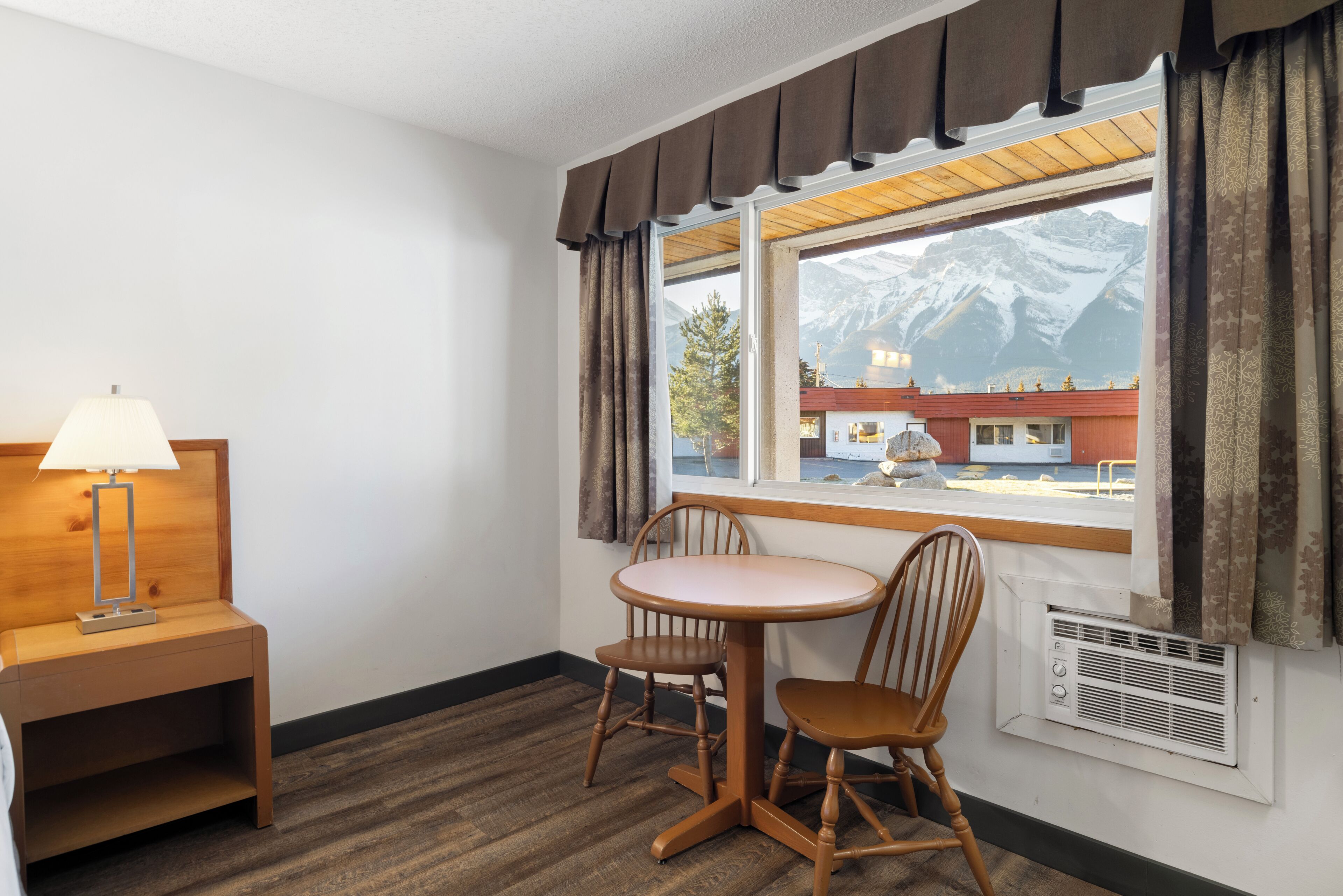 A hotel room with a wooden chair and table, a lamp, a window with curtains, and a view of a mountain.