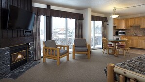 A cozy living room with a fireplace, two armchairs, a dining table with chairs, and a kitchen area with wooden cabinets and appliances. - Rocky Mountain Ski Lodge (Canmore)