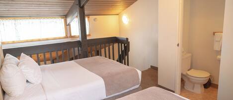 A bedroom with two beds, a wooden ceiling, a window with curtains, and a bathroom with a toilet and sink.
