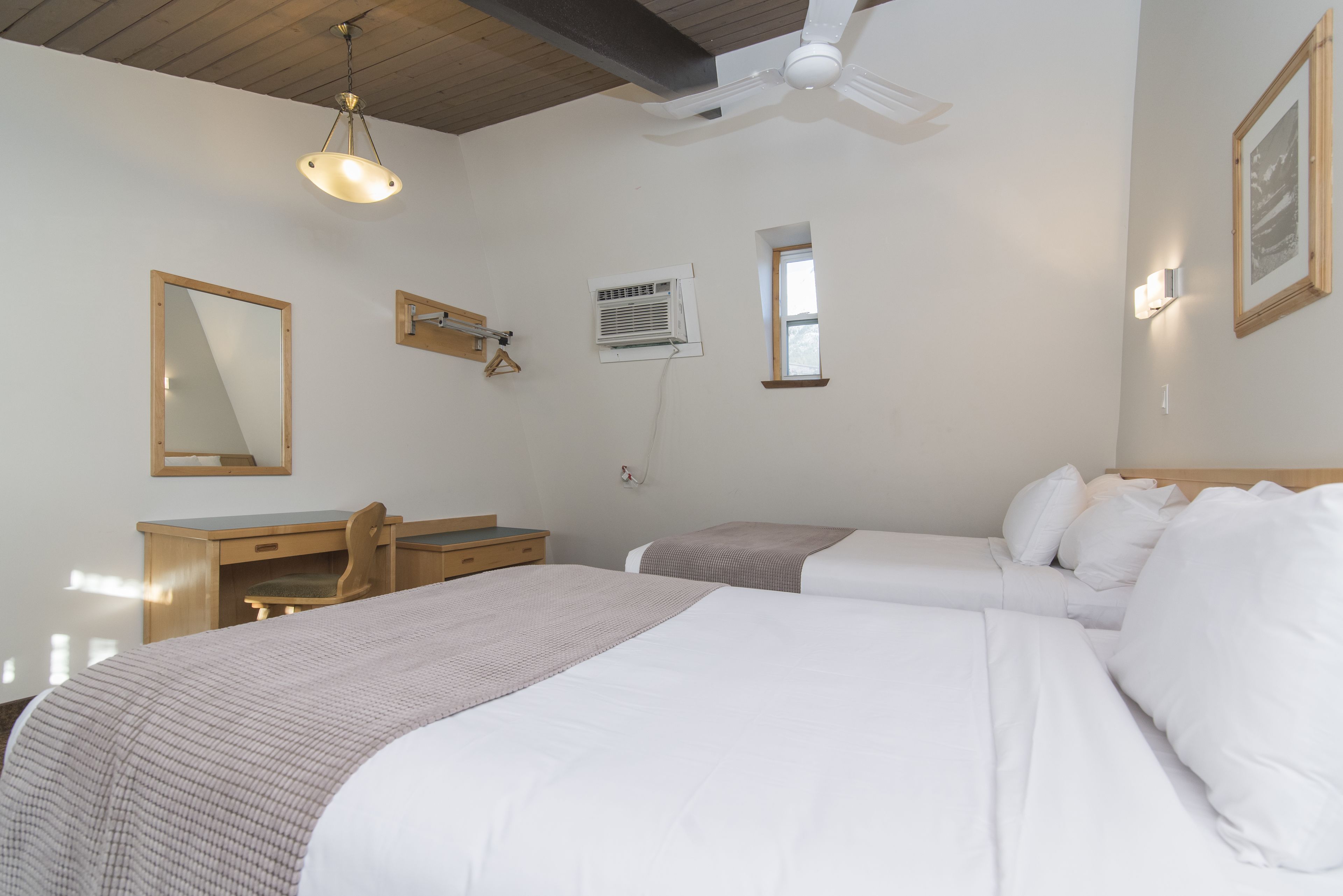 A hotel room with two beds, a desk, a chair, a ceiling fan, and a window.