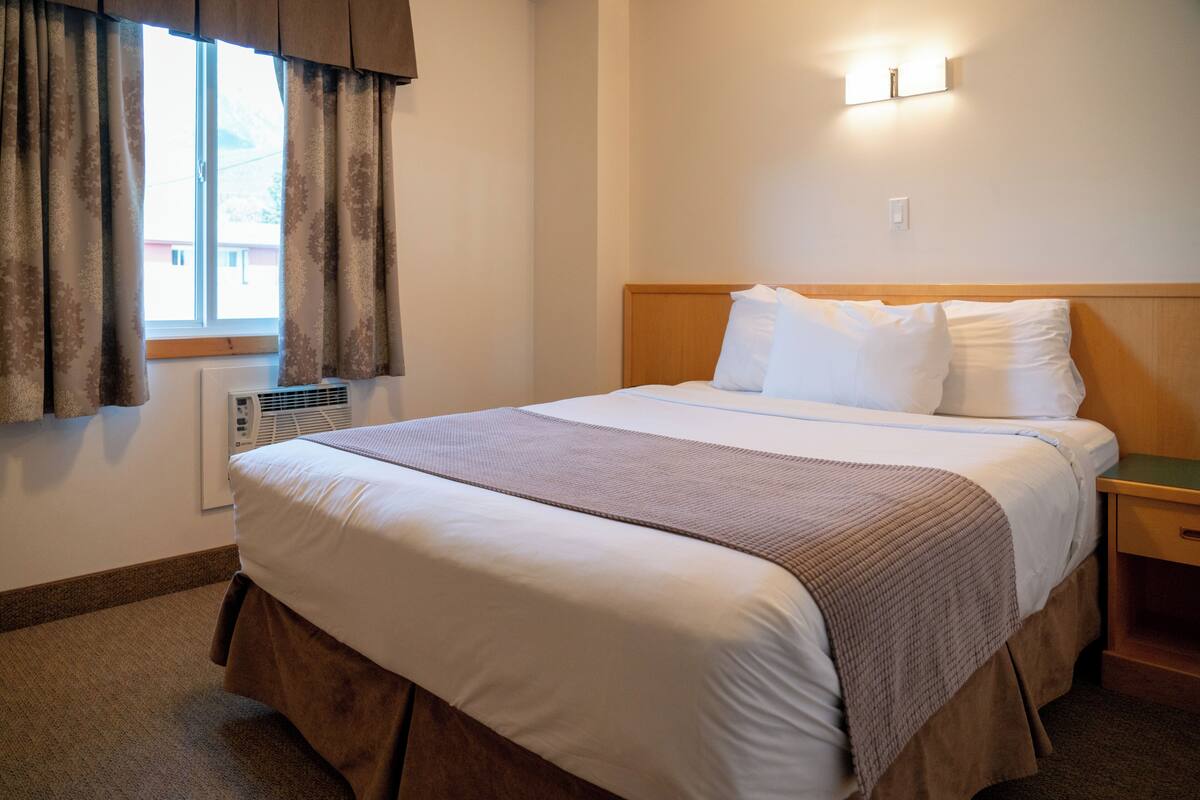 A hotel room with a bed, a window with curtains, a wall-mounted light, and a thermostat.